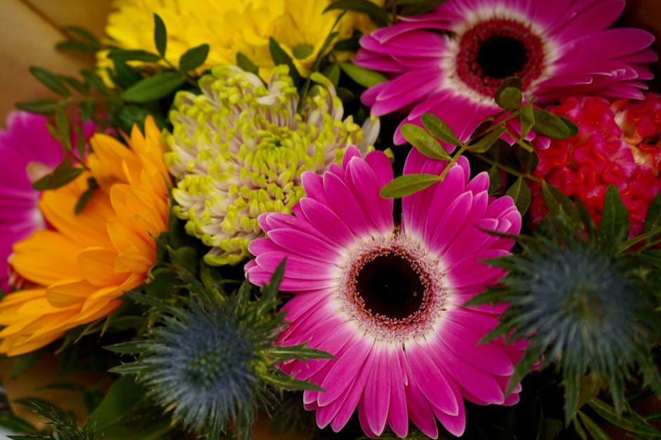 A vibrant bouquet of fresh flowers featuring large pink gerbera daisies with dark centers, bright yellow sunflower-like blooms, pale green chrysanthemums, and spiky blue thistles, all arranged with lu