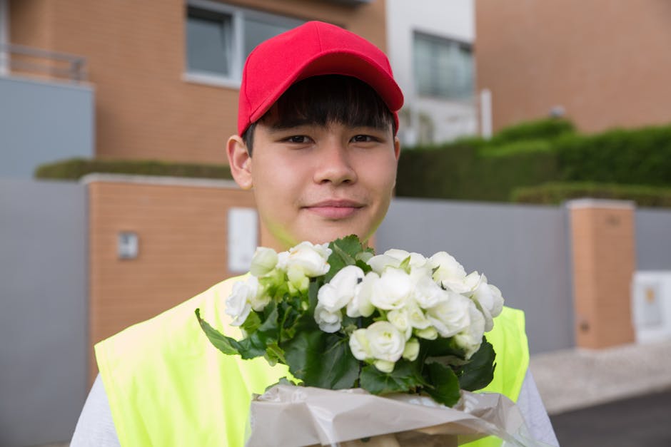 A young male flower delivery courier wearing a bright yellow high-visibility vest and a red cap, standing outdoors in front of modern residential buildings with greenery and brick walls. He is holding