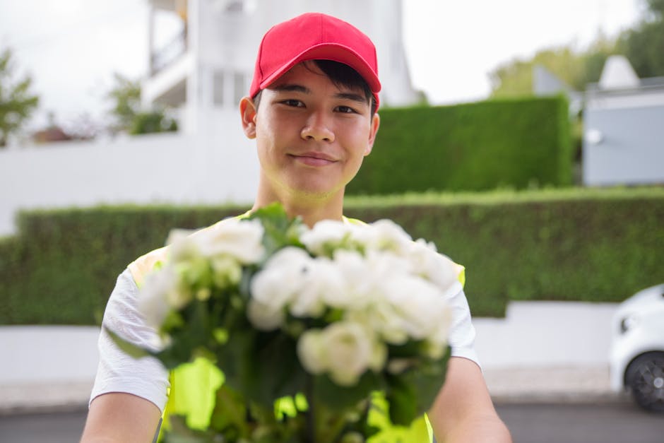 A delivery person wearing a red cap and a high-visibility vest holds out a bouquet of white roses and green foliage, with stems wrapped in clear paper. The individual is standing outdoors in a residen
