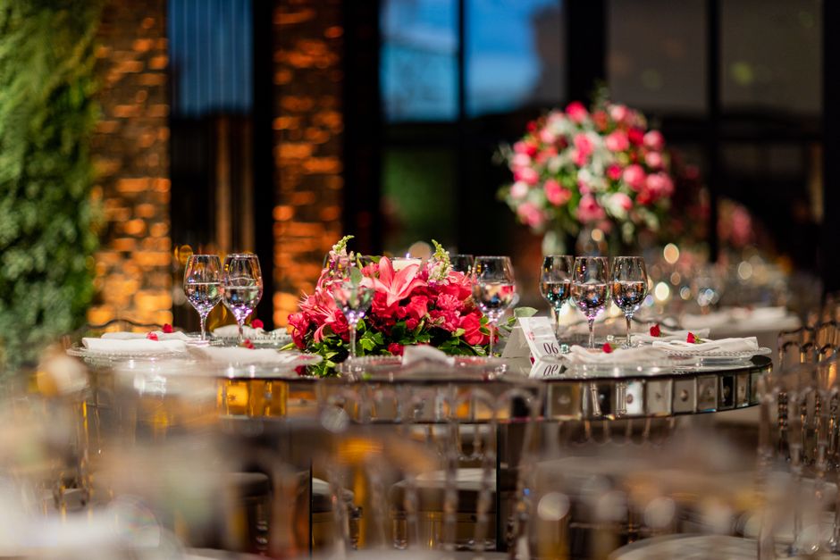 A beautifully arranged floral centerpiece featuring a lush bouquet of pink lilies, roses, and smaller filler flowers, all arranged on a round table set for a formal event. The bouquet is surrounded by