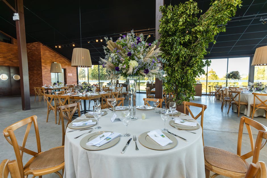 A round table in an elegant event venue arranged with a large floral centerpiece featuring white, purple, and lavender flowers, including roses and delphiniums, set in a tall glass vase. The table is 